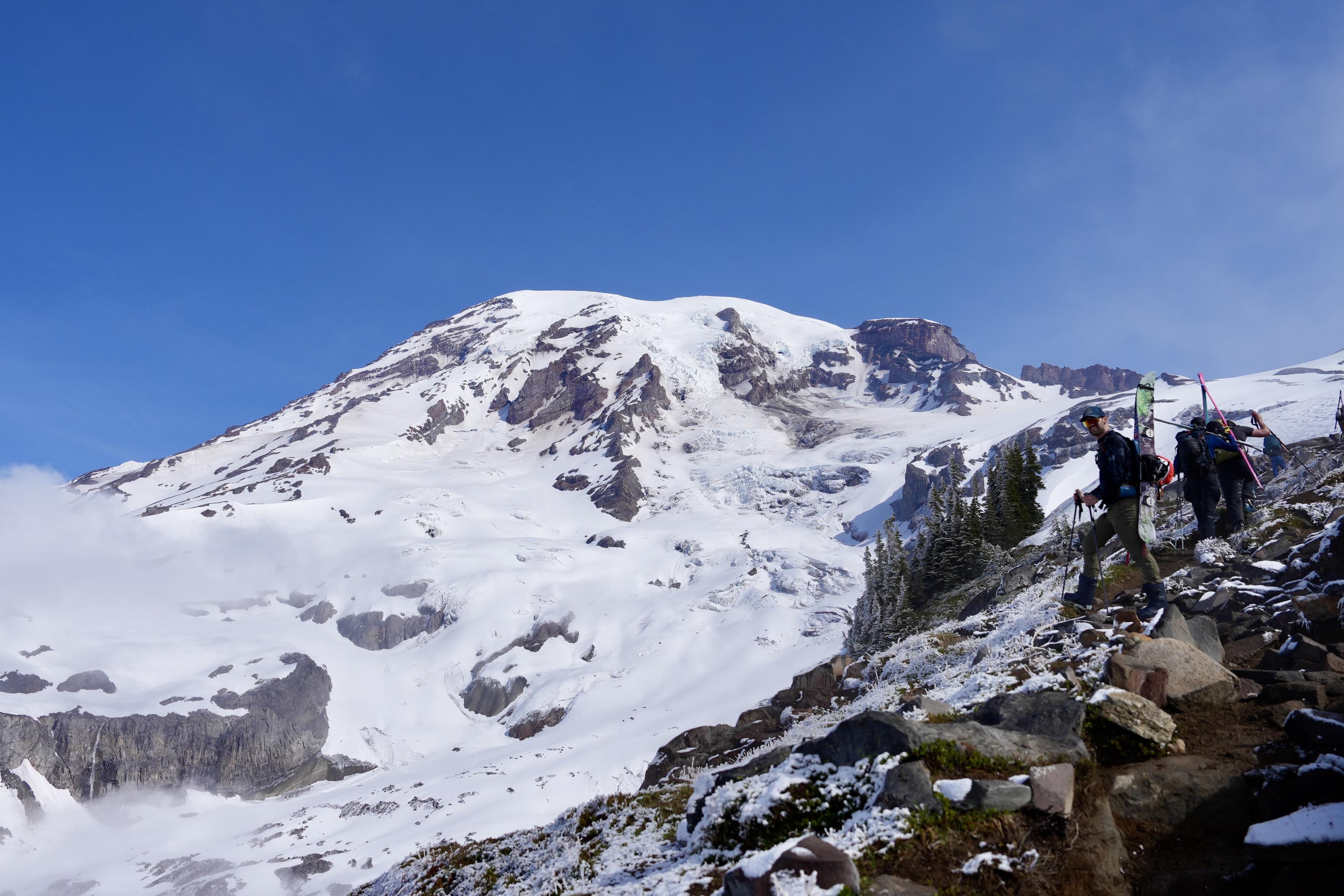 View from around Panorama Point looking up towards the summit of Mount Rainier. The mountain in the distance is almost completely covered in snow except for steep rocky areas. The immediate terrain is mostly free of snow and a clear trail can be seen. The sky is bright blue except for some thin wispy clouds. Three skiers carrying their skis in their arms or on their pack are in the foreground. An additional backpacker and skier can be seen further up the trail.