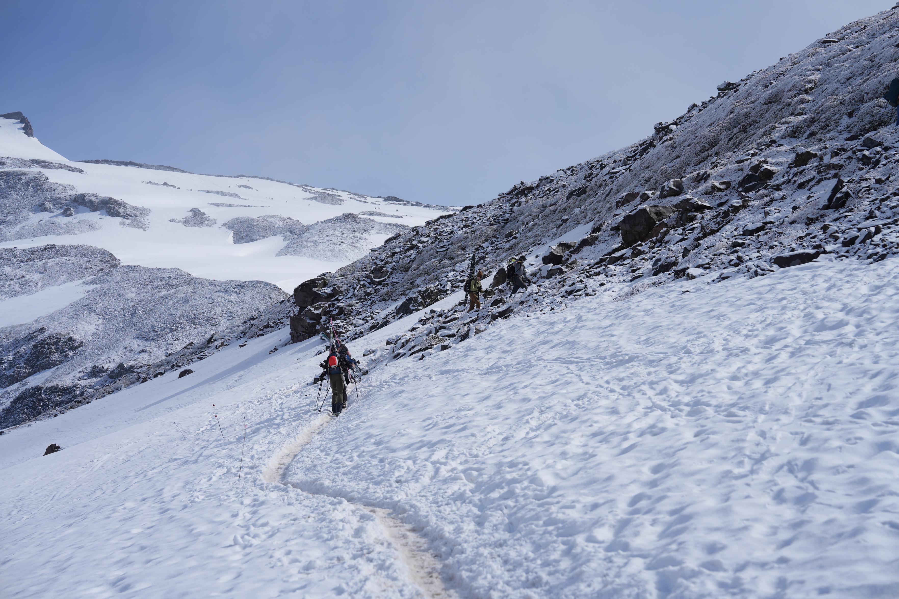 View from around Panorama Point looking up towards the Muir Snowfield. The snowfield in the distance and the immediate terrain is almost completely covered in snow except for steep rocky areas. Three skiers carrying their skis in their arms or on their pack are in the foreground hiking up the trail as it ascends a rocky area.