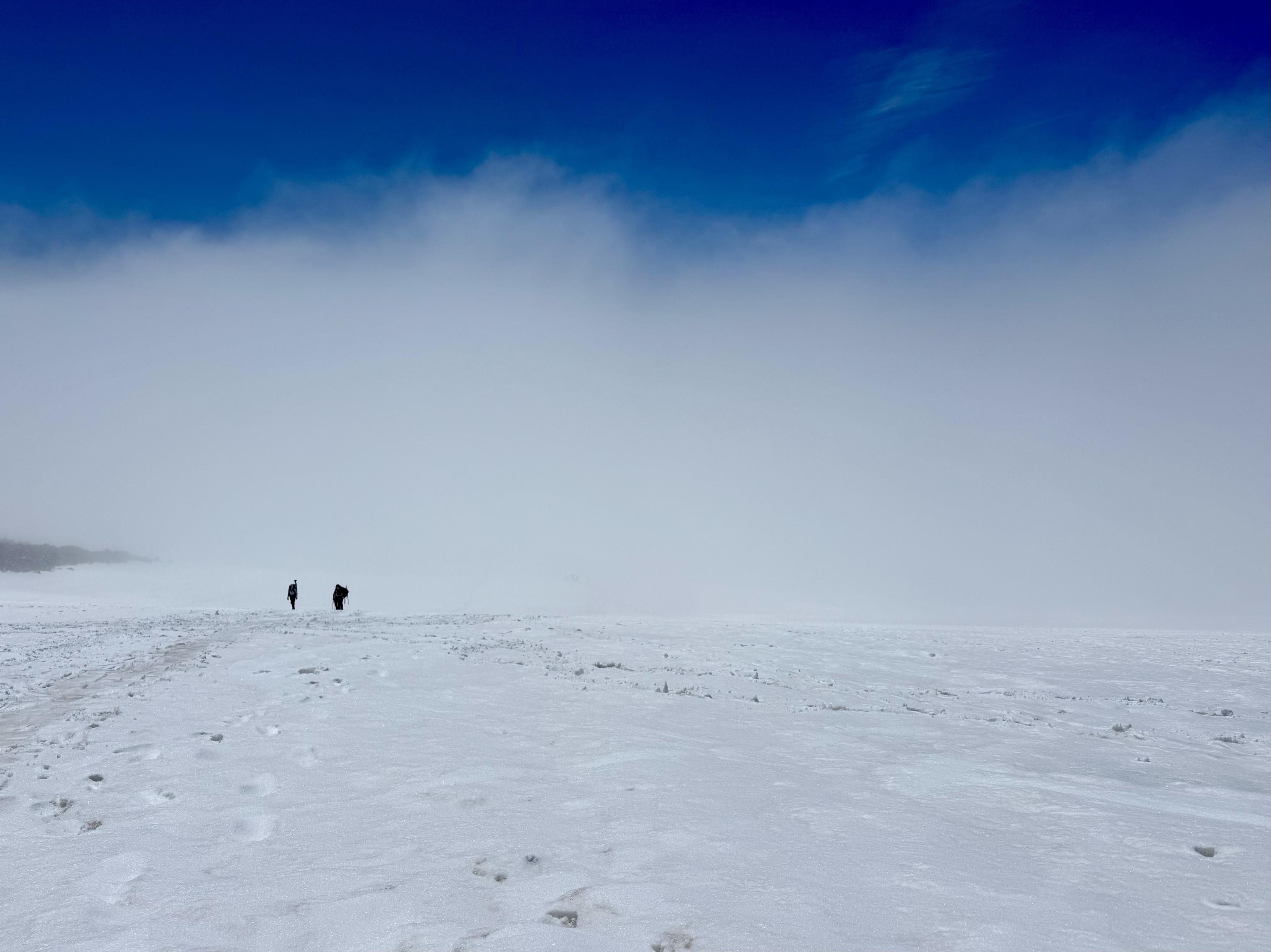 View from the middle of the Muir Snowfield looking back down the mountain. The immediate terrain is covered in snow full of holes from boots and tracks from skis. The view beyond the immediate snowfield is obscured clouds close to the terrain except for a bright blue strip of sky above. Three backpackers can be seen ascending the snowfield in front of the clouds.