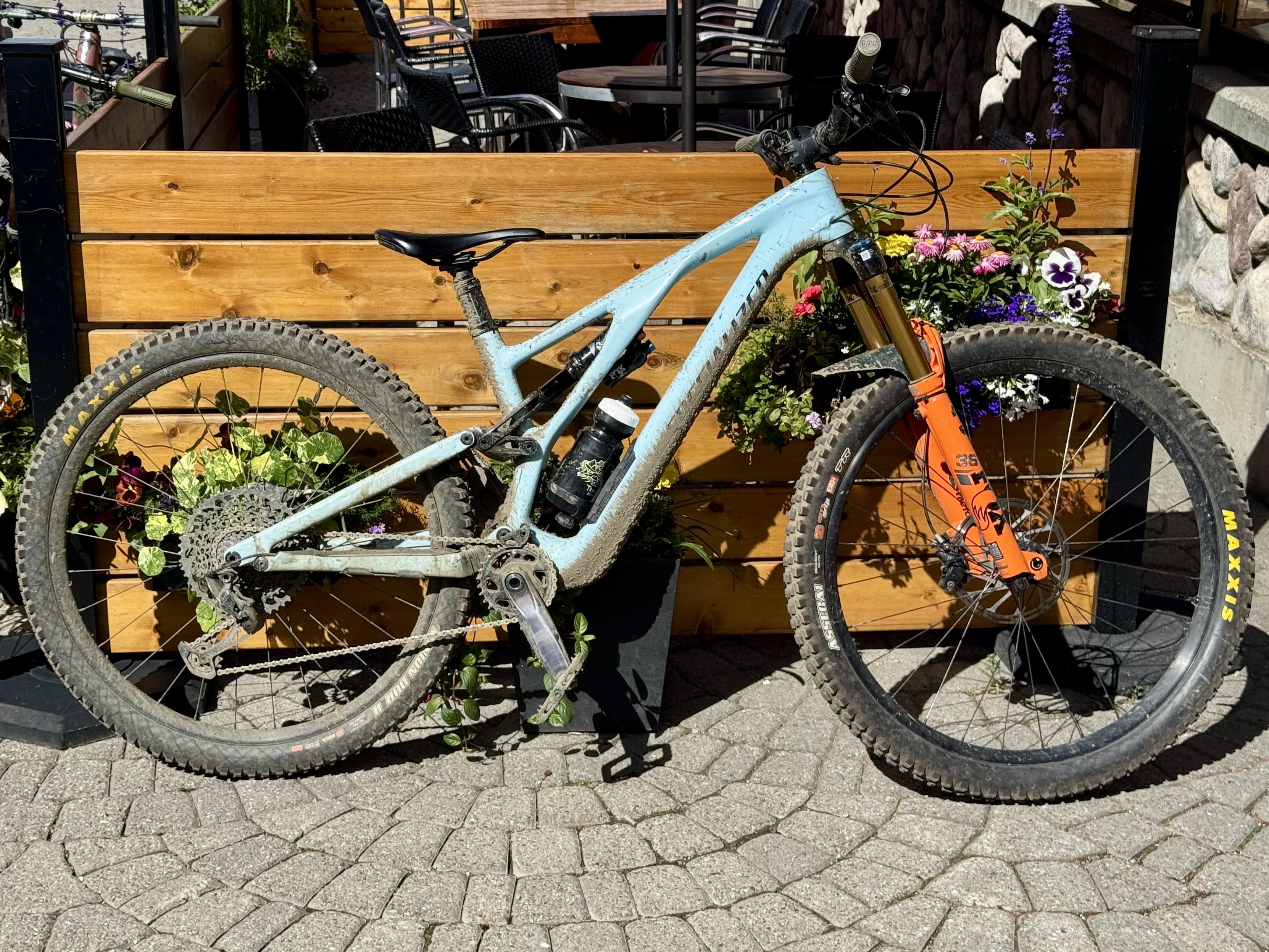 Photo of a mountain bike leaning against a low wooden fence with potted flowers In between. The bike frame is a light glossy blue while the front fork is orange. The frame is covered in mud kicked up from the tires.